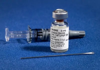 A two-pronged needle, a glass vial of smallpox vaccine, and a syringe sit on a blue surface.